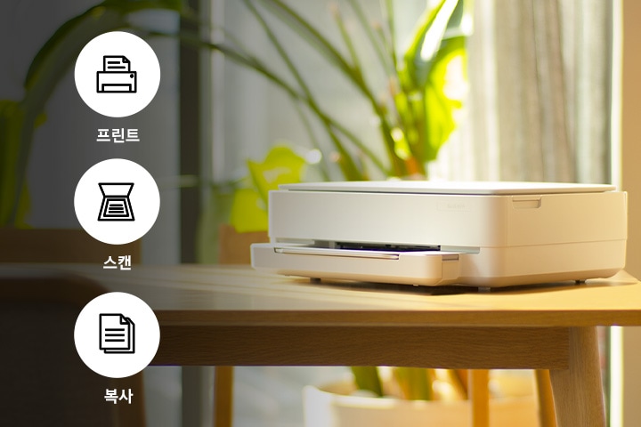 햇빛이 쏟아지는 창문 앞네 식물이 닜는 배경에 식탁 위에 제품이 왼쪽으로 20도 돌아간 이미지가 왼쪽에 배치되어 있는 사진입니다. 제품 이미지 왼쪽에는 프린터 텍스트와 프린터 이미지, 스캔 텍스트와 스캔 이미지, 복사 텍스트와 복사 아이콘이 왼쪽에서 오른쪽으로 나열되어 있는 이미지 입니다.