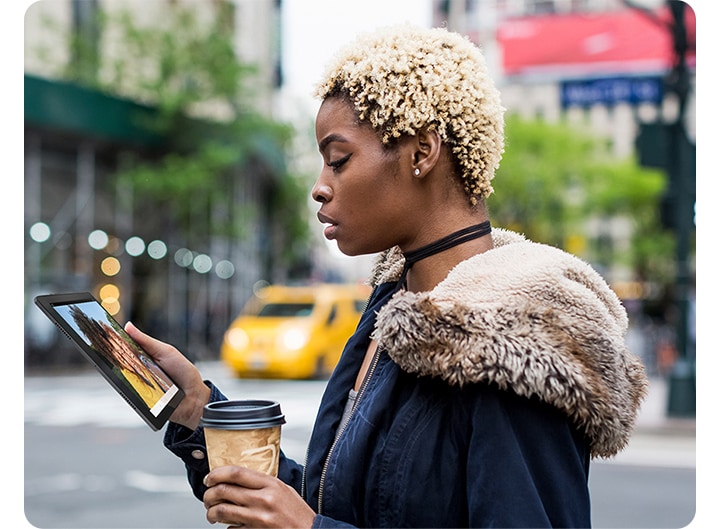 한 손에는 갤럭시 탭 A8 기기를, 다른 한 손에는 테이크아웃 음료를 들고 있는 여성이 시내 거리에서 화면을 보고 있습니다.