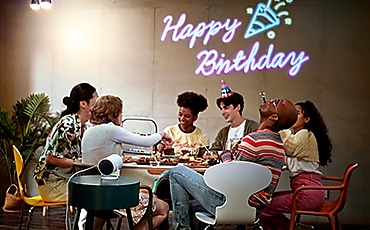 여성, 남성 사용자들이 테이블에 둘러앉아 있습니다. 벽면에는 생일축하 네온사인 화면이 보입니다.