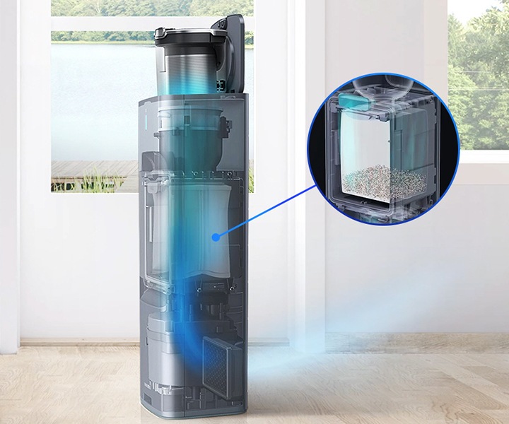 원목 마루가 깔린 거실 뒤에는 하얀 벽이 있고 벽 가운데와 오른쪽에는 호수가 보이는 창문이 있습니다. 창문 가장 왼쪽에는 흰색의 얇은 커튼이 드리워져 있습니다. 이미지 왼쪽 하단에는 원형 테이블이 보이고 이미지 가장 가운데 청정스테이션에 먼지통이 결합되어 있는 모습이 보입니다. 청정스테이션 내부는 투명하게 처리되어 안 쪽 내부 부품이 투명하게 보이는 상태로 가장 하단 청정스테이션용 배기필터 부분을 파란색 선으로 표시 후 확대하여 보여주소 있습니다. 