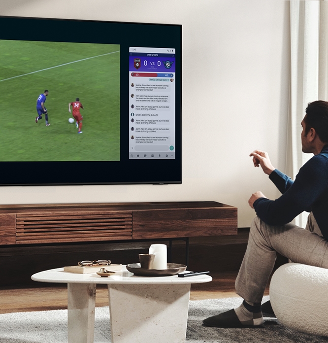 남성이 소파에 앉아 TV 를 보고 있습니다. TV 화면은 두개의 분할로 나뉘어 있습니다. 좌측에는 축구 중계화면이 보이고 우측에는 채팅창에 메세지들이 보이고 있습니다.