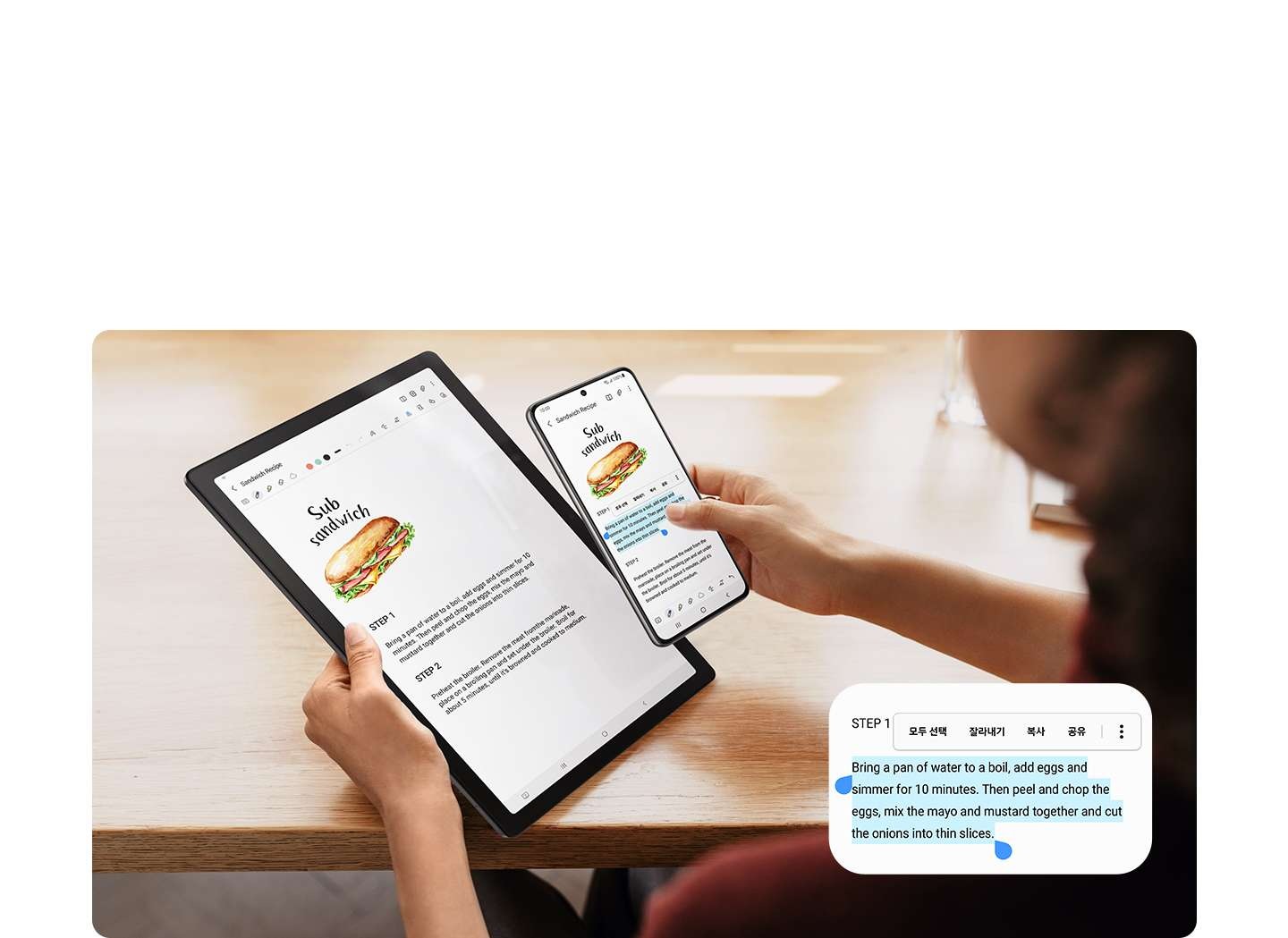한 손에는 갤럭시 탭 A8, 다른 한 손에는 갤럭시 스마트폰을 들고 있습니다. 스마트폰 화면에 텍스트가 선택되어 표시되고 복사 및 붙여넣기 옵션이 팝업되어 있습니다.