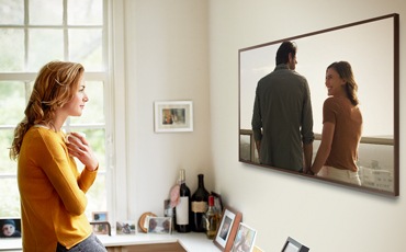 벽걸이 TV가 가로 모드로 설치되어 있고 사용자가 서서 TV를 바라보고 있습니다. 화면에는 남성과 여성이 보입니다.