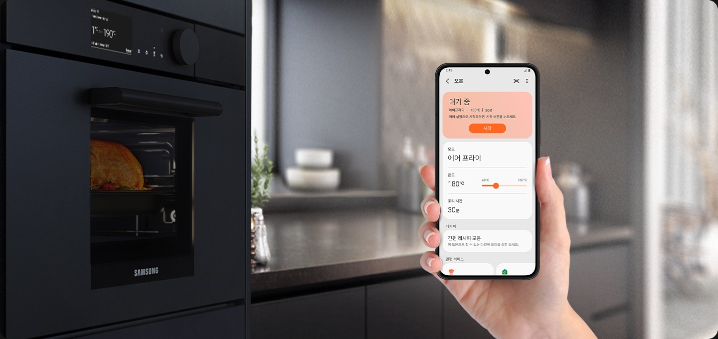 왼쪽에 빌트인 되어있는 오븐이있고, 오른쪽에는 핸드폰을 들고 있는 손이 있습니다. 핸드폰 디스플레이 안에는 스마트싱스 앱이 사용 되고 있고 맨 위에는 대기 중 문구와 함께 에어프라이 | 180도 | 30분 아래 설정으로 시작하려면, 시작 버튼을 눌러주세요. 시작 버튼이 활성화 되어있습니다. 그 아래에는 모드 > 에어 프라이, 온도 > 180도, 조리 시간 30분, 그 아래에 레시피 간편 레시피 모음 이 오븐으로 할 수 있는 다양한 요리를 살펴보세요. 문구가 있습니다.