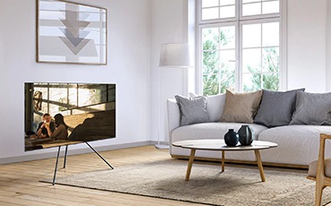 거실에 TV와 스탠드가 결합된 제품이 45도 각도로 놓여있습니다. 화면 안에는 인물들의 모습이 보입니다.