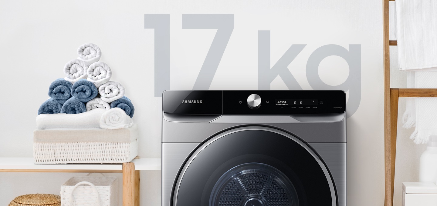 욕실로 보이는 공간에 삼성 그랑데 건조기 AI 제품이 놓여져 있습니다. 왼쪽 선반에는 수건이 쌓여져 있고 그 밑에는 바구니가 있습니다.