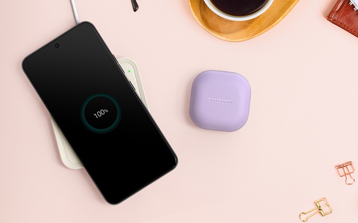 분홍색 배경에 커피와 공책 클립들이 놓여있고, 중앙에 갤럭시 버즈와 스마트폰이 있습니다. 스마트폰 아래에는 노티드 무선 충전기가 있습니다.