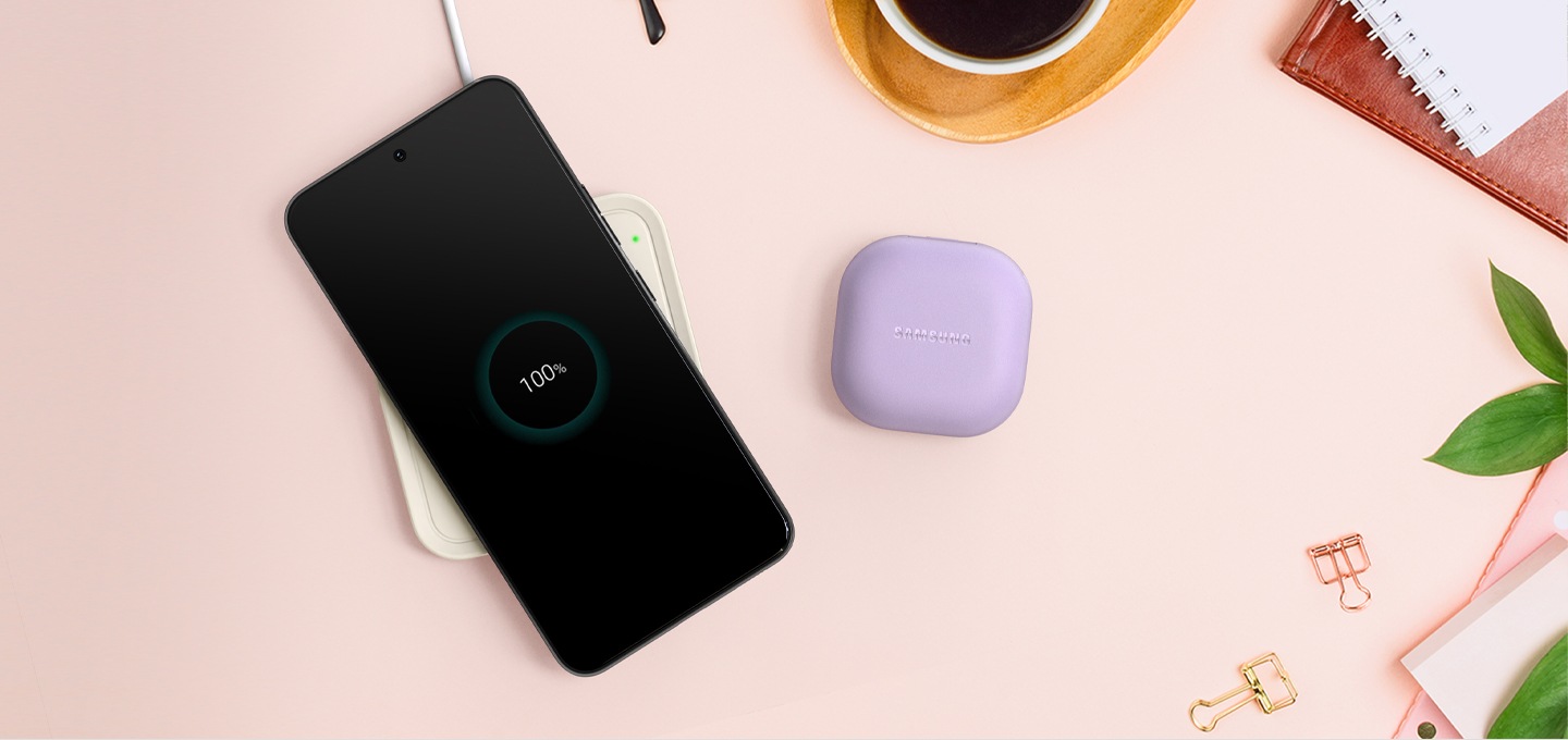 분홍색 배경에 커피와 공책 클립들이 놓여있고, 중앙에 갤럭시 버즈와 스마트폰이 있습니다. 스마트폰 아래에는 노티드 무선 충전기가 있습니다.