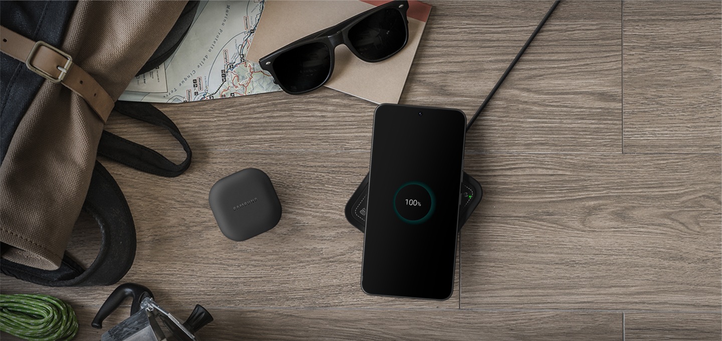 우드 테이블 위에 가방과 썬글라스, 지도, 공책이 놓여져 있고 중앙에 갤럭시 버즈와 갤럭시 스마트폰이 놓여져 있습니다. 스마트폰 아래에는 하이브로우 무선 충전기가 있습니다.