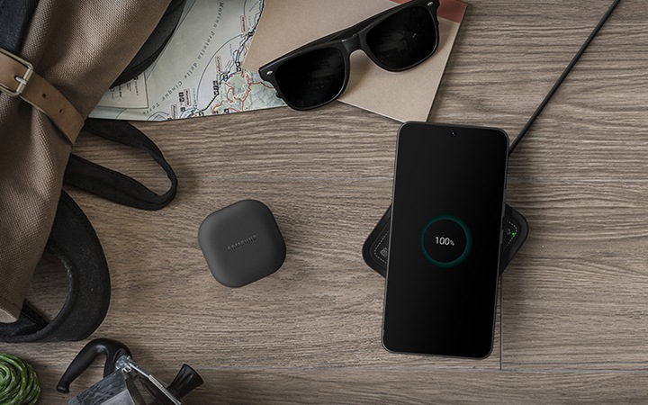 우드 테이블 위에 가방과 썬글라스, 지도, 공책이 놓여져 있고 중앙에 갤럭시 버즈와 갤럭시 스마트폰이 놓여져 있습니다. 스마트폰 아래에는 하이브로우 무선 충전기가 있습니다.