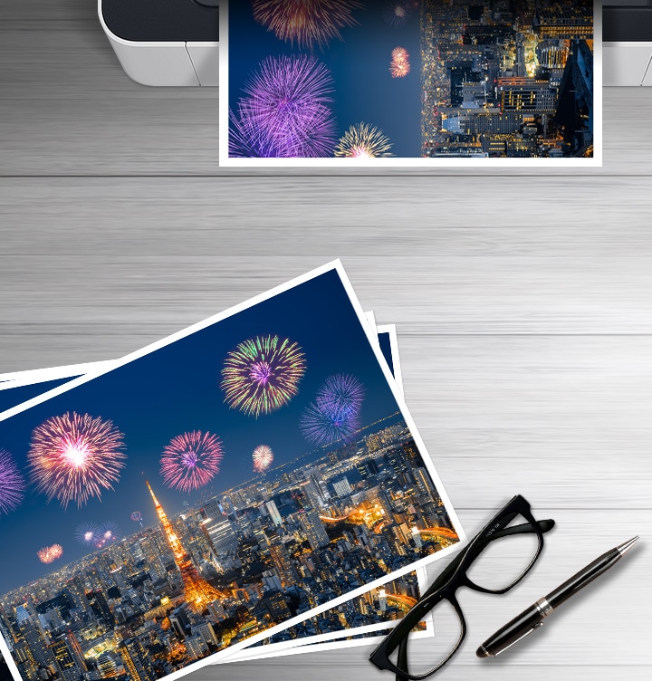 이미지 왼쪽 하단에는 폭죽놀이를 하고 있는 도시의 야경을 보여주는 이미지가 출력되어 있고 이미지 오른쪽 하단 부분에는 안경과 볼펜에 이미지가 있습니다. 이미지 오른쪽 상단부분에는 폭죽놀이를 하고 있는 도시의 야경을 출력하는 프린트기를 상단에서 바라본 이미지가 배치하여 있습니다.