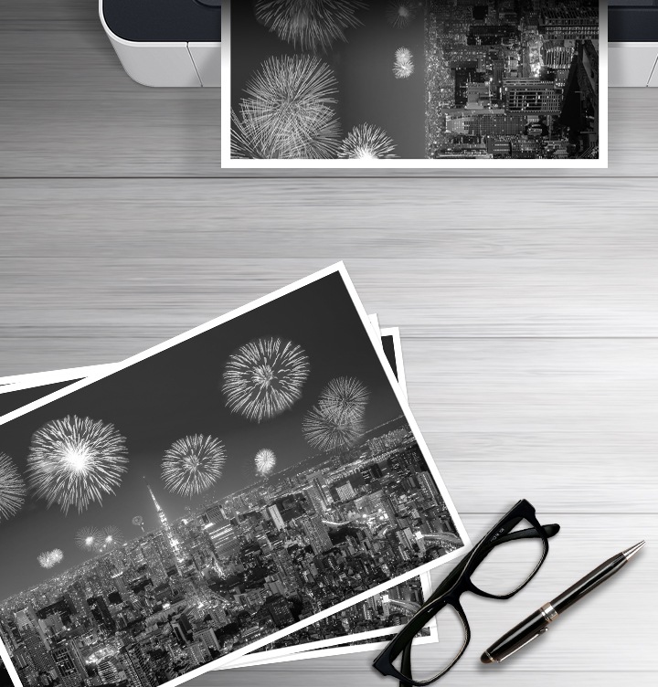 이미지 왼쪽 하단에는 폭죽놀이를 하고 있는 도시의 야경을 보여주는 이미지가 출력되어 있고 이미지 오른쪽 하단 부분에는 안경과 볼펜에 이미지가 있습니다. 이미지 오른쪽 상단부분에는 폭죽놀이를 하고 있는 도시의 야경을 출력하는 프린트기를 상단에서 바라본 이미지가 배치하여 있습니다.