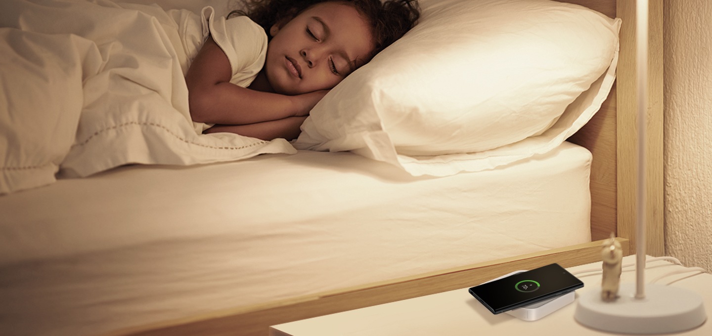 잠을 자고 있는 아이 옆에 테이블이 놓여져 있습니다. 그리고 그 테이블 위에 스마트싱스 스테이션이 있고 스마트싱스 스테이션 위에 갤럭시 휴대폰이 놓여져 있습니다. 휴대폰은 충전중입니다.