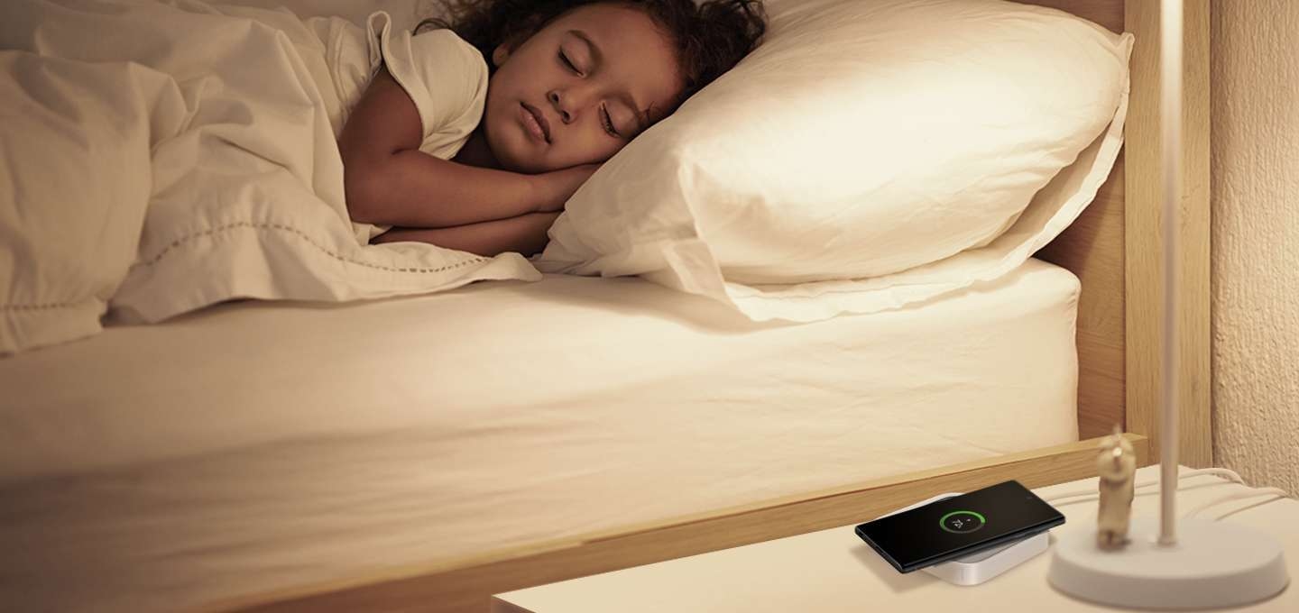 잠을 자고 있는 아이 옆에 테이블이 놓여져 있습니다. 그리고 그 테이블 위에 스마트싱스 스테이션이 있고 스마트싱스 스테이션 위에 갤럭시 휴대폰이 놓여져 있습니다. 휴대폰은 충전중입니다.