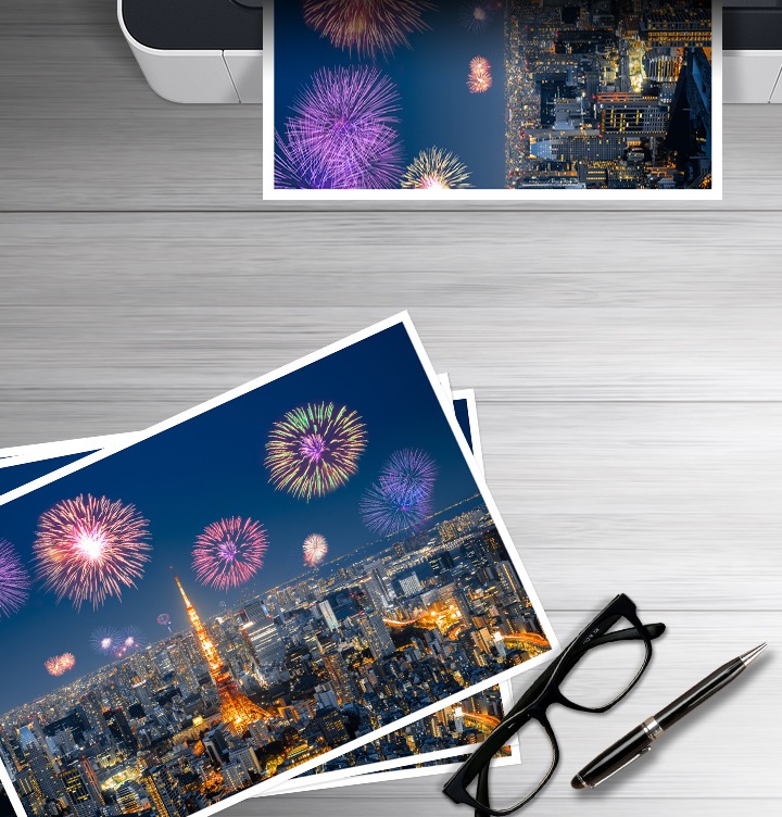 이미지 왼쪽 하단에는 폭죽놀이를 하고 있는 도시의 야경을 보여주는 이미지가 출력되어 있고 이미지 오른쪽 하단 부분에는 안경과 볼펜에 이미지가 있습니다. 이미지 오른쪽 상단부분에는 폭죽놀이를 하고 있는 도시의 야경을 출력하는 프린트기를 상단에서 바라본 이미지가 배치하여 있습니다.