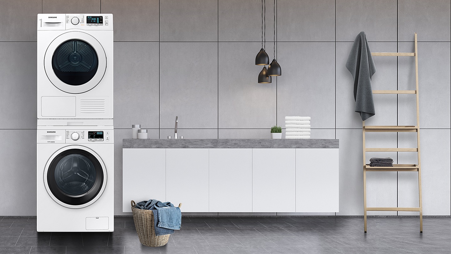 그레이 타일로 인테리어 되어있는 넓은 다용도실이 보이며, 좌측엔 비스포크 그랑데 AI 세탁기 건조기가 건조기 상단키트를 사용해 하나로 되어 있는 모습이 보이며, 가운데에 수납공간이 하단에 함께있는 세면대가 보이며 세면대 위 좌측엔 세탁세제 통이 보이고 우측엔 개어져 있는 하얀색 수건 6개가 보여지고 수건 우측엔 작은 흰 화분이 보여집니다.  세면대 앞엔 라탄의 빨래통과 안에 청바지 빨래가 들어있으며, 우측엔 사다리가 있으며 사딜에 상단에 수건이 걸려있고 두번째 칸에 수건이 개어져 있습니다.