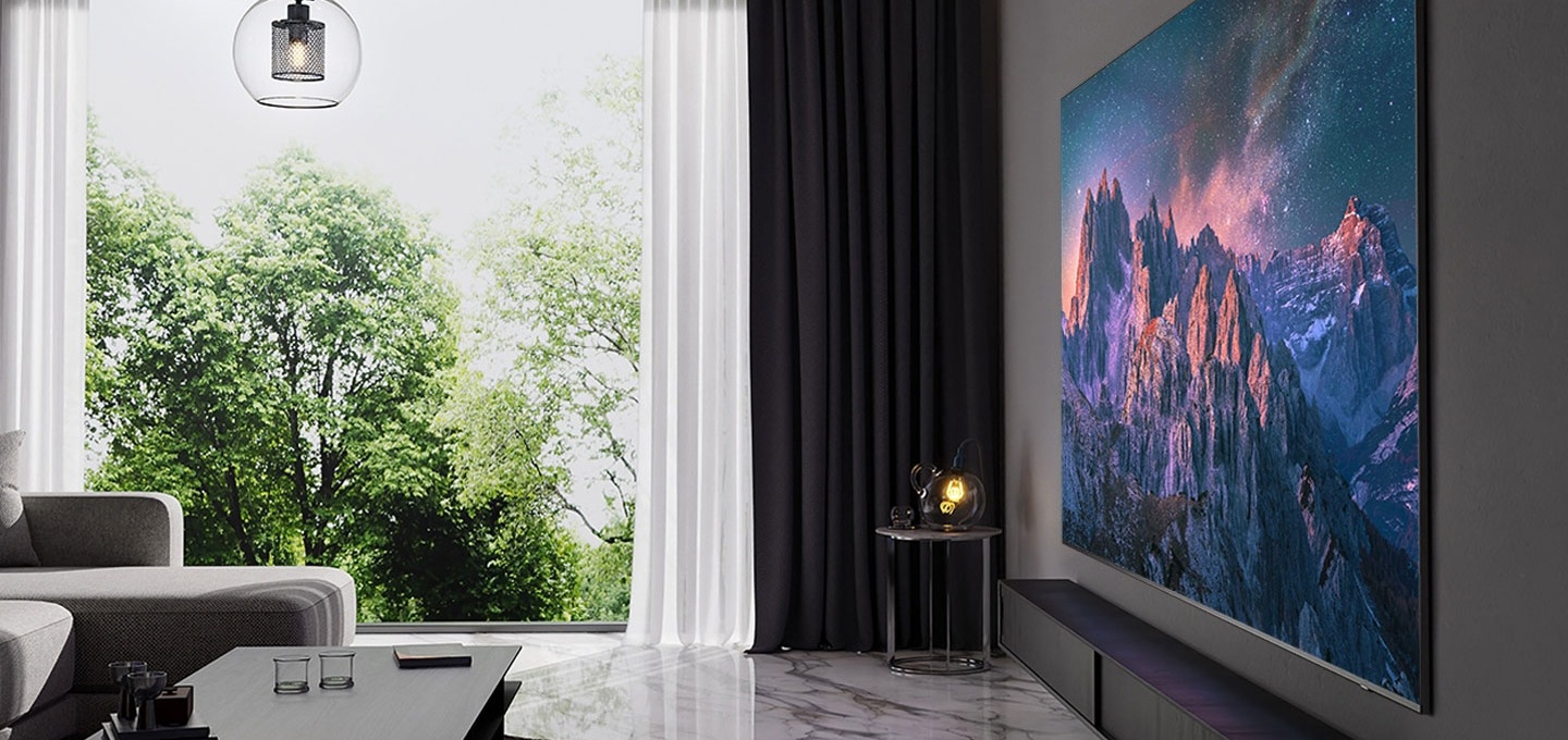 거실 벽에에 초대형 TV가 설치되어 있습니다. 스크린에는 산들의 모습이 보입니다. TV 왼쪽에 있는 창문은 열려 있으며 나무가 보입니다. TV 앞에는 쇼파와 티 테이블이 있습니다.