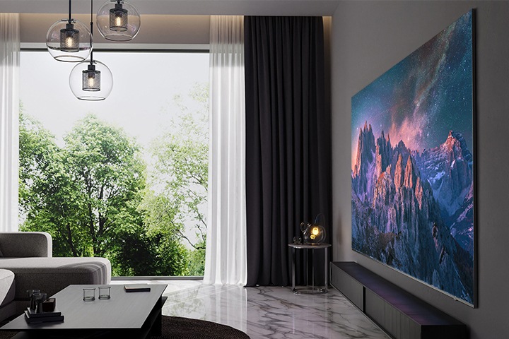 거실 벽에에 초대형 TV가 설치되어 있습니다. 스크린에는 산들의 모습이 보입니다. TV 왼쪽에 있는 창문은 열려 있으며 나무가 보입니다. TV 앞에는 쇼파와 티 테이블이 있습니다.