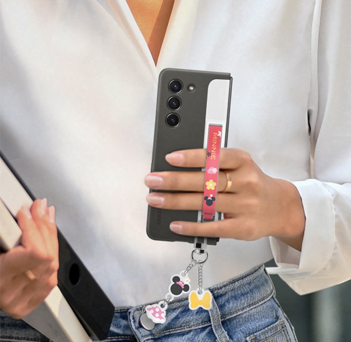 세미 정장 차림의 젊은 여성이 갤럭시 Z 폴드 스탠딩 케이스 with 스트랩 제품에서 스트랩 액세사리를 장착한 갤럭시 Z 폴드를 손가락을 넣어 들고 있습니다. 케이스 아래에는 다양한 악세서리가 달려 있습니다.