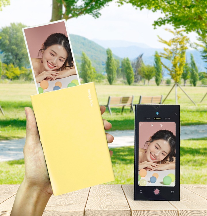 공원 배경에서 모바일 포토 프린터 옐루우 제품을 사용하여 사진을 프린트 하고 있는 이미지