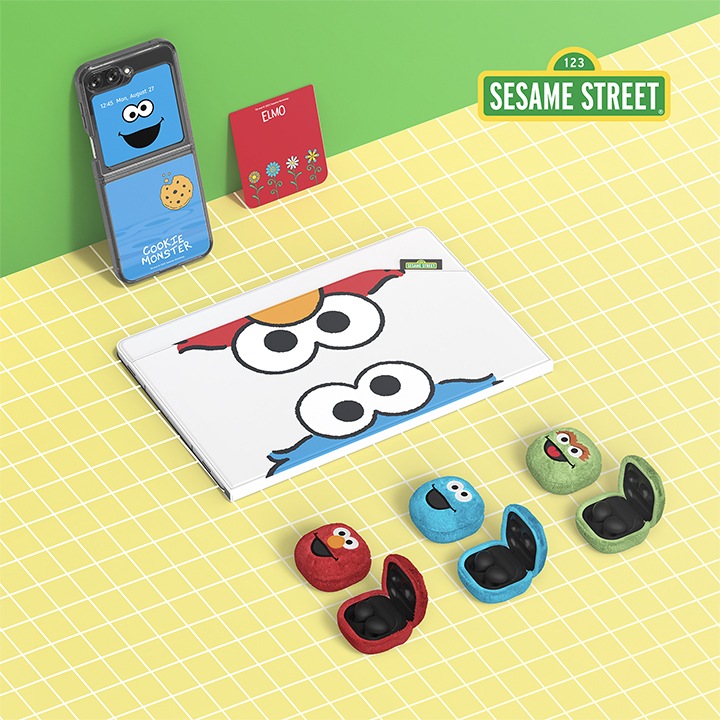 노란색 타일 패턴 바닥과 녹색의 벽이 배경으로 보여지며 오른쪽 상단에 세서미 스트리트 로고가 보여집니다. 세서미 스트리트 케이스를 장착한 갤럭시 Z 플립과 세서미 스트리트 플립수트 카드가 벽에 기대어져 보여지며, 세서미 스트리스 앤벨롭 커버를 장착 하고 있는 갤럭시 탭이 보여지며 아래에 3개의 갤럭시 버즈가 세서미 스트리트 버즈케이스 (오스카), 세서미 스트리트 버즈케이스 (쿠키몬스터), 세서미 스트리트 버즈케이스 (엘모) 순서로 보여지고 있습니다.