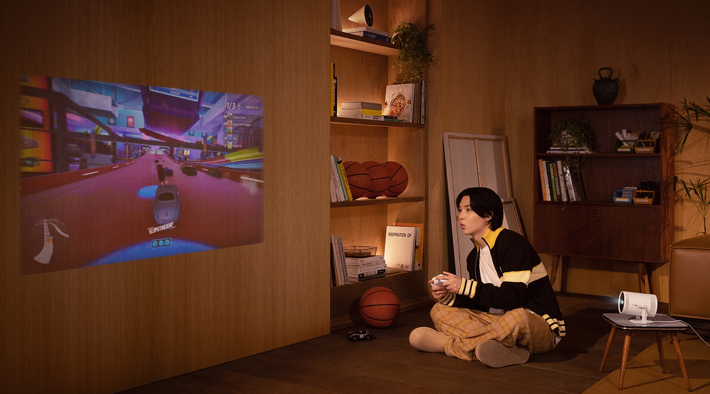실내 거실 바닥에 앉아 게임을 하고 SUGA 모습입니다. 옆 테이블에 놓여진 the freestyle이 벽을 향해 켜져 있고 화면에는 게임 장면이 보입니다.