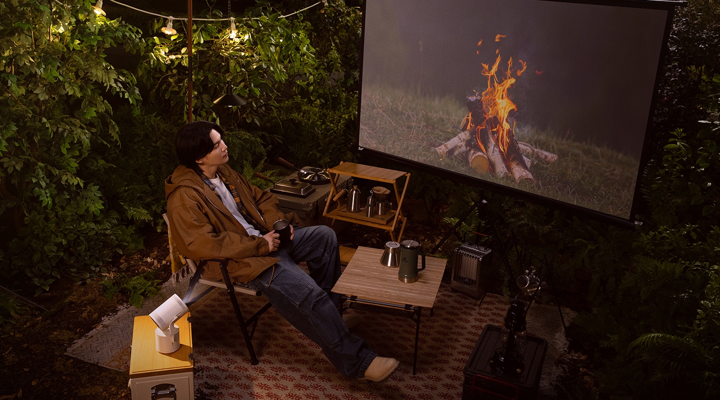 숲풀 사이 캠핑 의자에 앉아 삼각대에 설치된 스크린을 보고 있는 SUGA 모습입니다. 테이블에 놓인 the freestyle이 스크린을 비추고 화면에는 모닥불 영상이 나오고 있습니다.