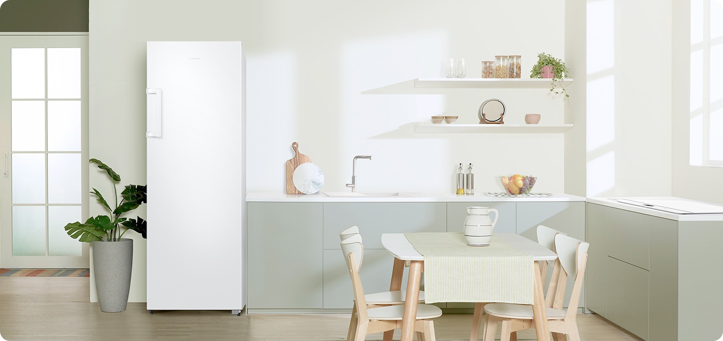 화이트/그린톤 주방 인테리어컷 입니다. 좌측에 벽 뒷쪽에 다용도실 유리문이 있습니다. 주방에는 좌측에 큰 화분이 있고 화이트 색상 냉동고가 설치되어 있습니다. 냉동고 우측에는 그린톤의 싱크대가 있고 앞쪽에는 4인용 식탁이 놓여있습니다.