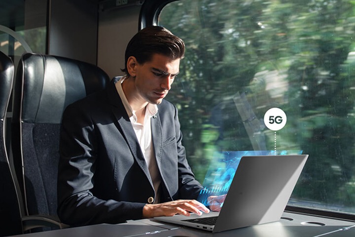 한 남성이 움직이는 기차의 창가 좌석에서 갤럭시 북3 Go를 사용하고 있습니다. 5G 기호가 갤럭시 북3 Go 위에 표시되어 있습니다.
