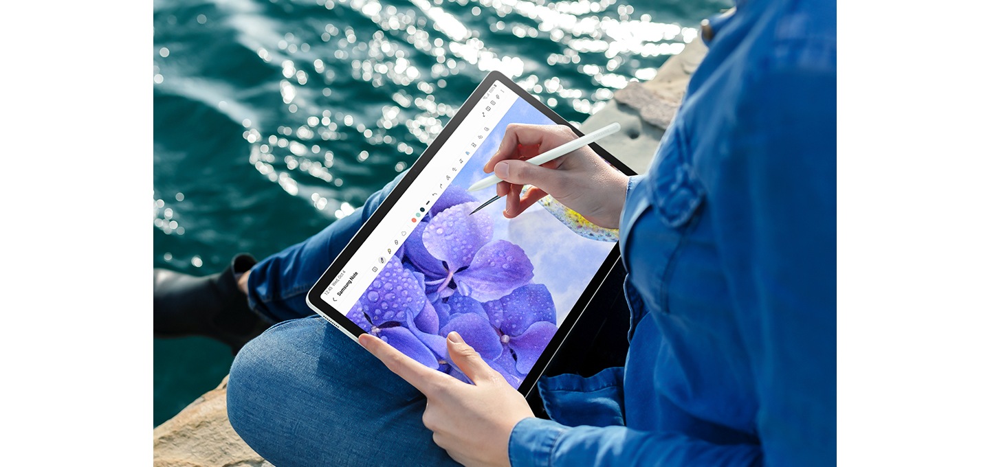 한 사람이 물가에 앉아 갤럭시 탭 S9 FE+를 가로 모드로 들고 있습니다. 화면에 열려 있는 Samsung Notes 앱에 S펜을 사용해 꽃 그림을 그리고 있습니다.