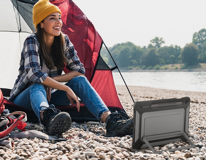이미지 오른쪽 부분에는 강과 숲이 보이고 있고 이미지 왼쪽 부분에는 빨간 색상의 텐트를 치고 그 옆에는 빨간 가방을 바닥에 내려놓은 여성이 텐드 앞에 앉아 있습니다. 여성은 노란 비니를 쓰고 체크무늬의 남방과 청바지를 입고 양쪽 무릎에  팔꿈치를 기대어 앉아있습니다. 여성에 앞에는 갤럭시 탭 S9 FE 및 S9 FE+ 제품에 아웃도어 커버를 장착한 제품의 정면 이미지가 보이고 있습니다. 