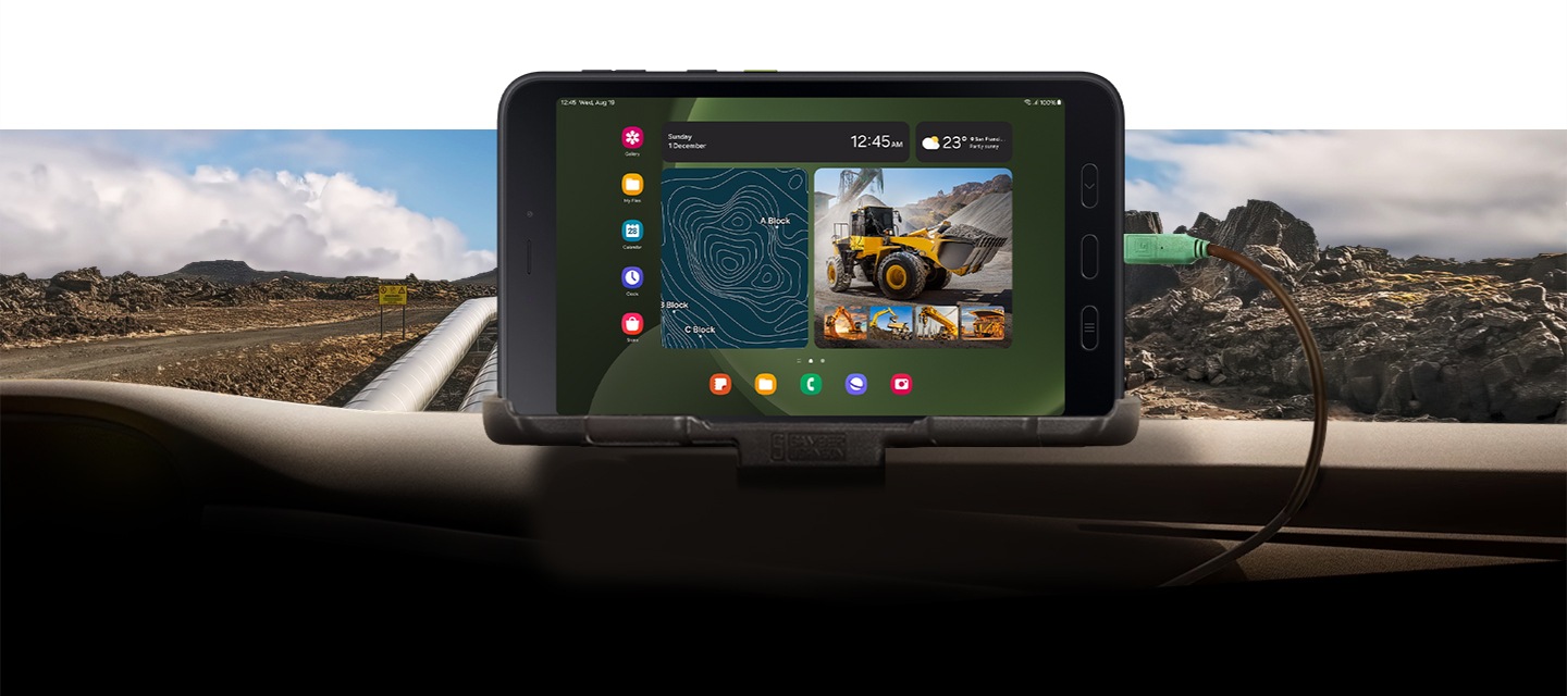 갤럭시 탭 액티브5가 자동차 대시보드에 장착되어 있습니다. 화면에 GPS 지도와 중장비 차량 이미지를 표시하고 있습니다. 기기는 자동차와 케이블로 연결되어 있고 배경에는 거친 바위가 많은 지형이 표시되어 있습니다.