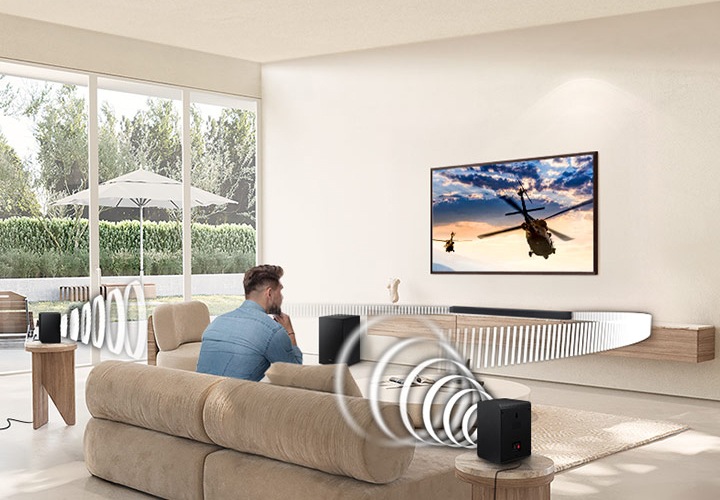 남성이 자리에 앉아 TV화면을 바라보고 있습니다. TV 화면에는 헬리콥터가 날아가는 보습이 보입니다. 사운드바와 리어 스피커에서는 음향효과가 나오고 있습니다.