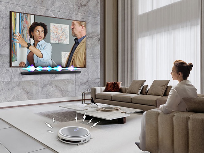 거실에서 여성이 TV를 보고 있습니다. TV 화면에는 드라마에 한 장면이 나오고 있습니다. TV 하단에는 사운드바가 설치되어 있고 TV와 사운드바 사이에는 음향효과가 있습니다. 거실 바닥에는 무선 로봇 청소기가 움직이고 있습니다.