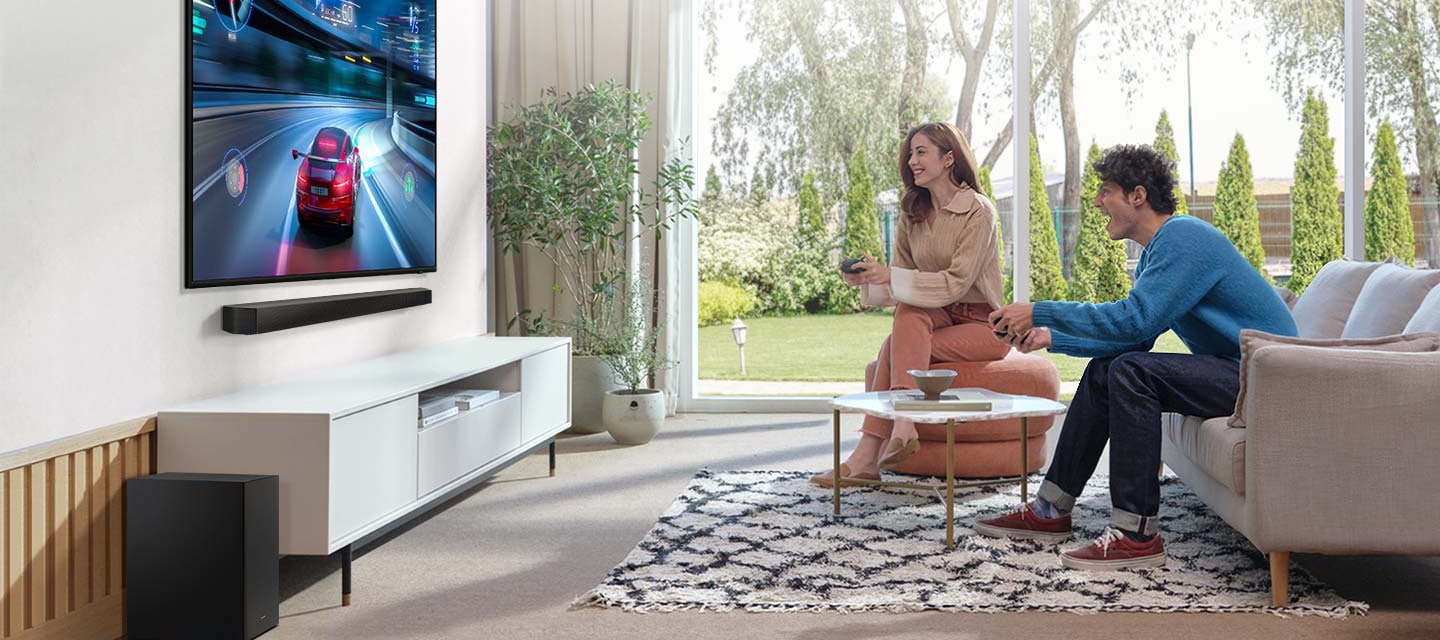 거실 벽면에 TV가 설치되어 있으며, TV 하단에는 장식장이 있고 그 위에 사운드바가 설치되어 있습니다. TV 화면에는 게임이 플레이되고 있으며, TV 정면 쇼파에서는 두명의 남녀가 게임을 즐기고 있는 모습입니다.