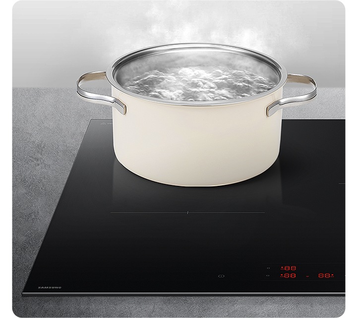 진한 대리석 상판 위에 삼성 인덕션이 설치되어 있고 그 위에 크림 컬러의 냄비가 올려져 있습니다. 냄비 안에는 물이 가득 들어 있고 수증기가 모락모락 올라오며 끓고 있습니다.