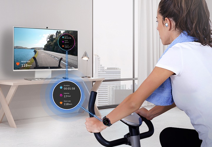 한 여성이 갤럭시 워치와 버즈를 착용한 상태로 헬스 사이클을 타고 있습니다. 스마트 모니터에는 운동 콘텐츠가 띄워져 있으며 모니터와 여성 사이에는 갤럭시 워치의 화면이 띄워져 있습니다.