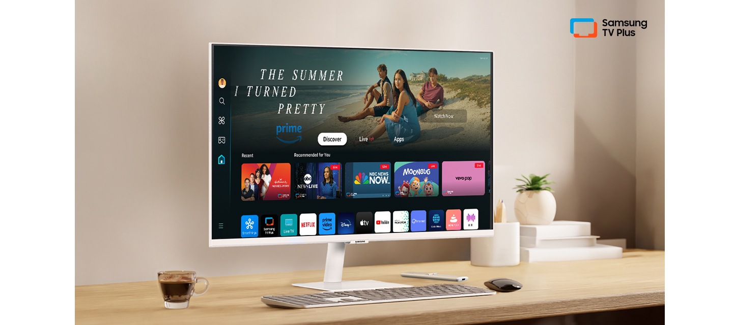 책상 위에 모니터가 있습니다. 화면 안에는 다양한 OTT 앱과 연결할 수 있는 홈 UI가 띄워져 있습니다. 오른쪽 상단에는 Samsung TV Plus 로고가 있습니다.