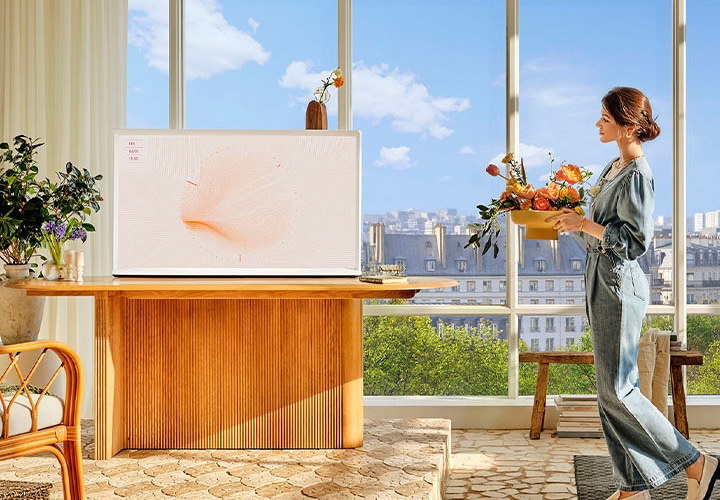 테이블 위에 TV가 설치되어 있습니다. TV 왼쪽에는 화병과 식물이 보이며, 오른쪽에는 여성이 꽃을 들고 있습니다.