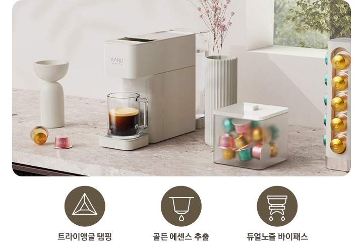 좌측에는 아이콘과 문구가 나와있습니다. 삼각형 아이콘과 트라이앵글 탬핑 문구, 캡슐에서 커피가 추출되는 아이콘과 골든 에센스 추출 문구, 커피머신에서 양쪽 노즐로 커피가 추출되는 아이콘과 듀얼노즐 바이패스 문구가 나와있습니다. 문구 우측에는 대리석 상판 위에 화병 등 오브제가 놓여있고 카누 캡슐커피머신 바리스타 브리즈 크림 화이트 컬러가 놓여있습니다. 머신 위 머그잔에는 아메키라노가 담겨있고 우측에 카누 전용 캡슐이 담긴 하얀색 반투명 상자가 놓여있습니다.