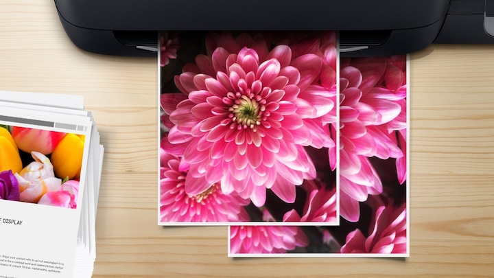 나무로 된 책상을 위에서 바라본 배경에 이미지 중간에는 꽃 이미지를 출력하고 있는 이미지와 이미지 왼쪽 하단에는 이미 출력된 출력물을 쌓아 놓은 이미지가 있습니다. 
