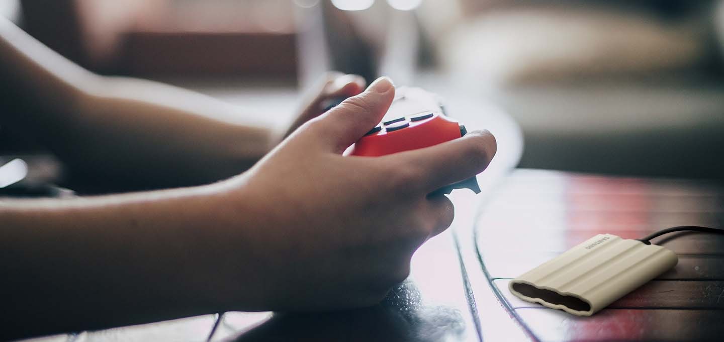 햇빛이 들어오는 실내에서 이미지 오른쪽에 외장 SSD 제품이 케이블로 게임에 연결되어 있는 사진입니다. 이미지 왼쪽에는 조이스틱을 조작하고 있는 사람의 손 부분이 확대외어 있습니다.