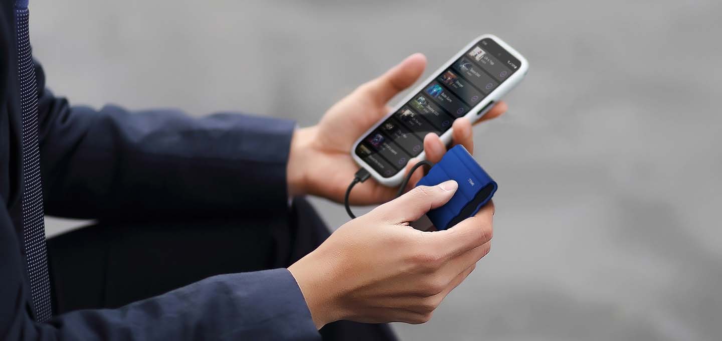 야외에서 외장 SSD 제품을 케이블을 이용하여 핸드폰에 연결하여 사용하는 남색 셔츠를 입은 남성의 손부분을 확대한 이미지 입니다. 