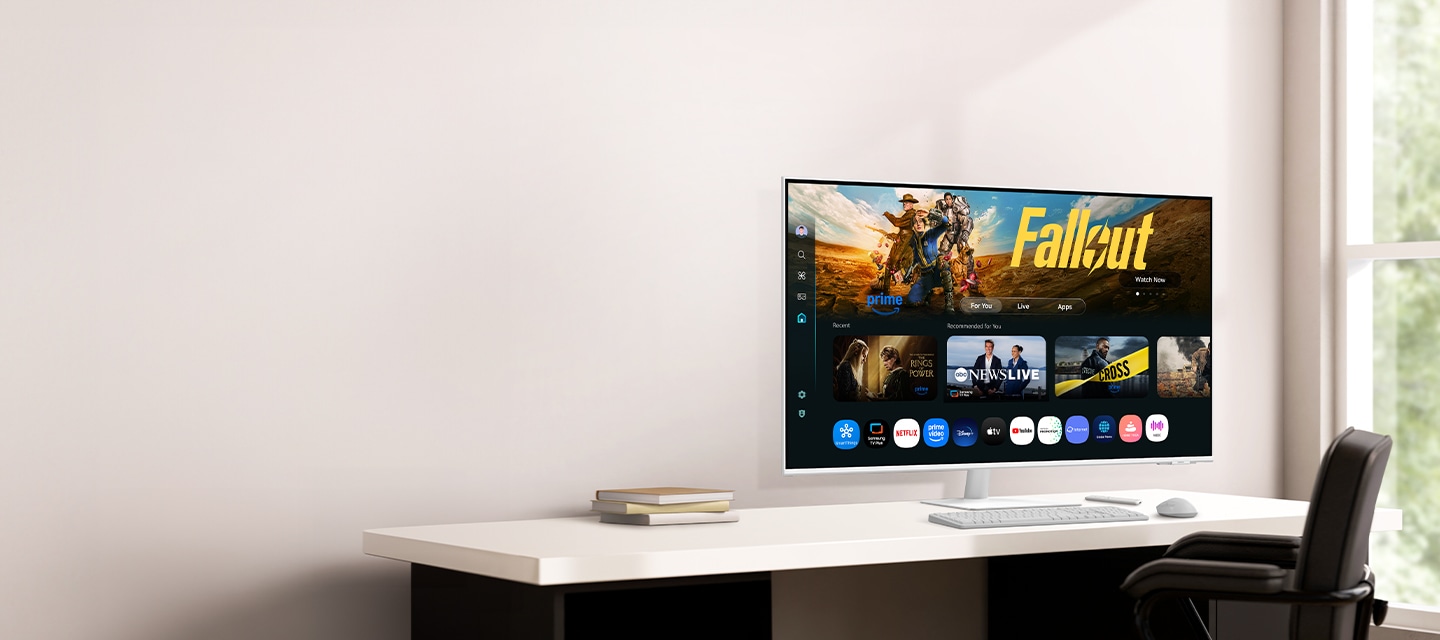 화이트 톤의 벽과 테이블에 삼성 스마트 모니터 M7,키보드, 마우스가 측면으로 배치되어 있으며, 화면에는 다양한 콘텐츠와 앱이 정렬된 스마트 TV 앱 페이지를  표시하고 있습니다.