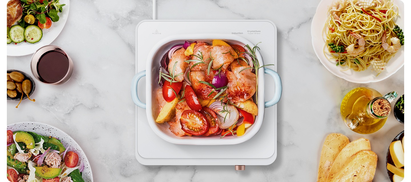 더 플레이트가 올려진 테이블이 탑 뷰로 확대되어 보입니다. 더 플레이트 1구 가운데에는 하얀색 냄비 안에 음식물이 조리되고 있고, 왼쪽부터 시계 방향으로 샐러그, 올리브, 오이와 야채, 파스타 오일소스, 바게트 빵이 보입니다.