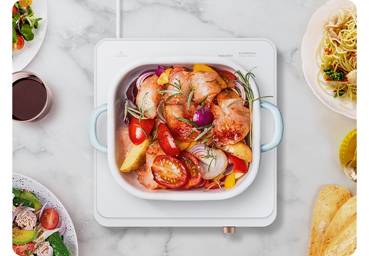 더 플레이트가 올려진 테이블이 탑 뷰로 확대되어 보입니다. 더 플레이트 1구 가운데에는 하얀색 냄비 안에 음식물이 조리되고 있고, 왼쪽부터 시계 방향으로 샐러그, 올리브, 오이와 야채, 파스타 오일소스, 바게트 빵이 보입니다.