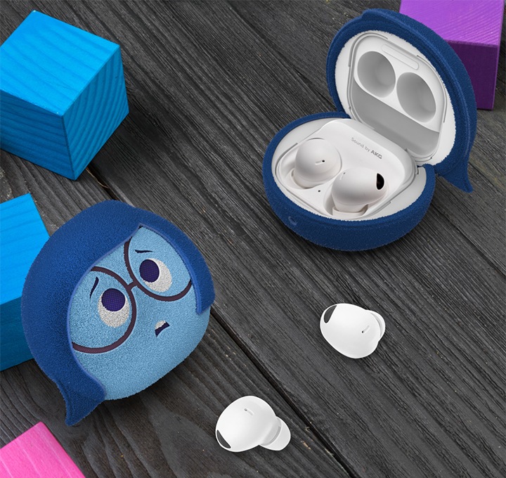 진한 나무 배경에 갤럭시 버즈 인사이드 아웃2 슬픔이 케이스를 장착하여 파란색 나무 블록에 기대어 놓은 정면 이미지와 갤럭시 버즈 인사이드 아웃2 슬픔이 케이스를 장착 후 열어 놓은 이미지가 왼쪽 하단에서 오른쪽 상단으로 배치되어 있습니다.