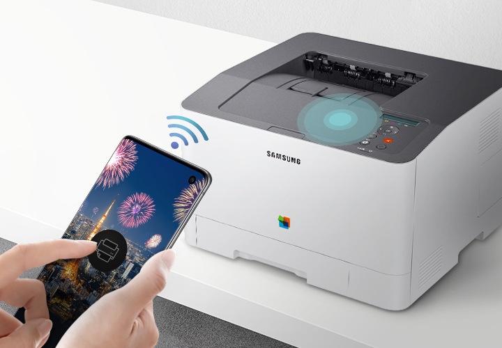 하얀색 책상 위에 제품을 오른쪽 위에서 내려본 이미지가 중간에 배치하여 있고 이미지 왼쪽 부분에 스마트폰 화면에 프린터 아이콘을 누르고 있는 사람의 손이 배치하여 있습니다.