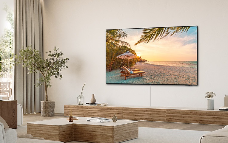 벽에 TV가 설치되어 있고, 화면에서는 해변이 보입니다. TV 하단에는 서랍장이 있으며 그 위에는 화병과 장식품이 놓여 있습니다. TV 앞에는 테이블과 소파가 놓여 있습니다.