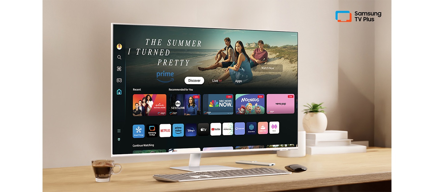 책상 위에 모니터가 있습니다. 화면 안에는 다양한 OTT 앱과 연결할 수 있는 홈 UI가 띄워져 있습니다. 오른쪽 상단에는 Samsung TV Plus 로고가 있습니다.